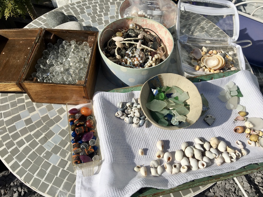 A studio table arranged with shells, sea glass, beads, discarded jewellery and crystal stones for mixed-media artwork.