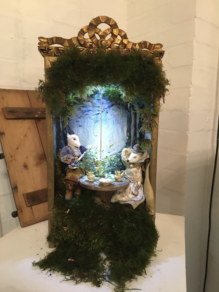 A lit moss-lined miniature theatre with two fox figures seated around a small table.