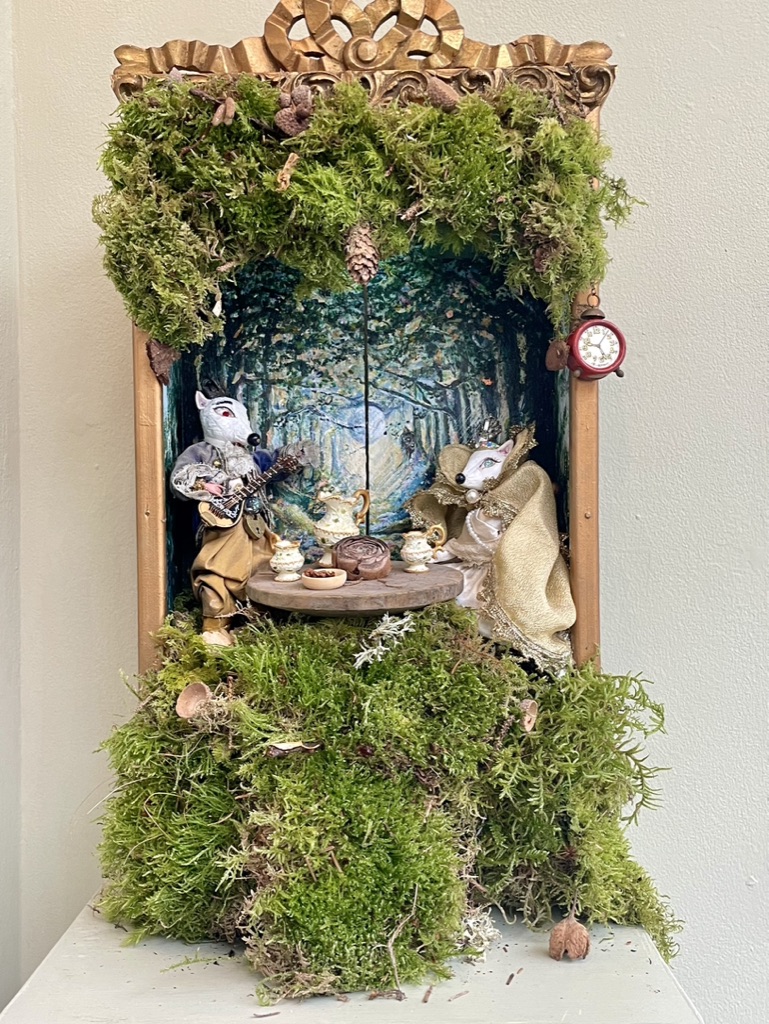 Moss-covered miniature theatre with fox figures, a tea table and a small red clock hanging at the side.