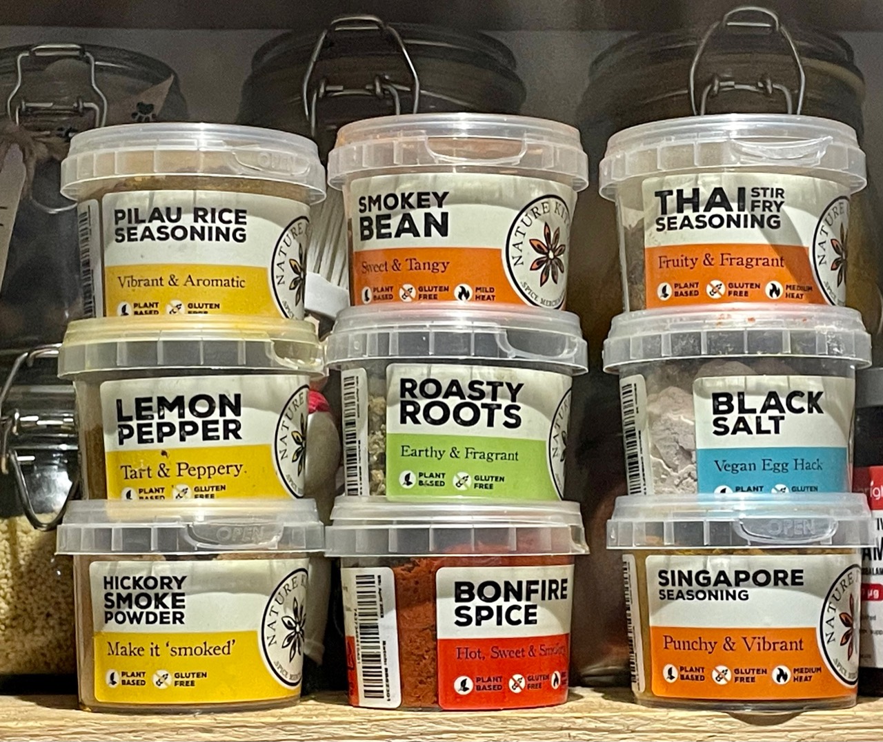 A pantry shelf filled with plant-based seasoning pots including smoky bean, Thai stir fry, lemon pepper, black salt and Singapore seasoning.