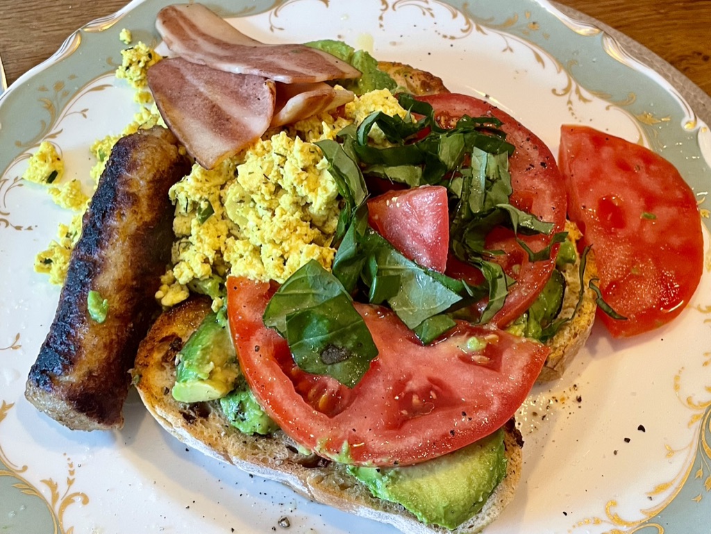Dirty vegan breakfast with sausage, vegan bacon, scrambled tofu, avocado and beef tomato on brown sourdough.