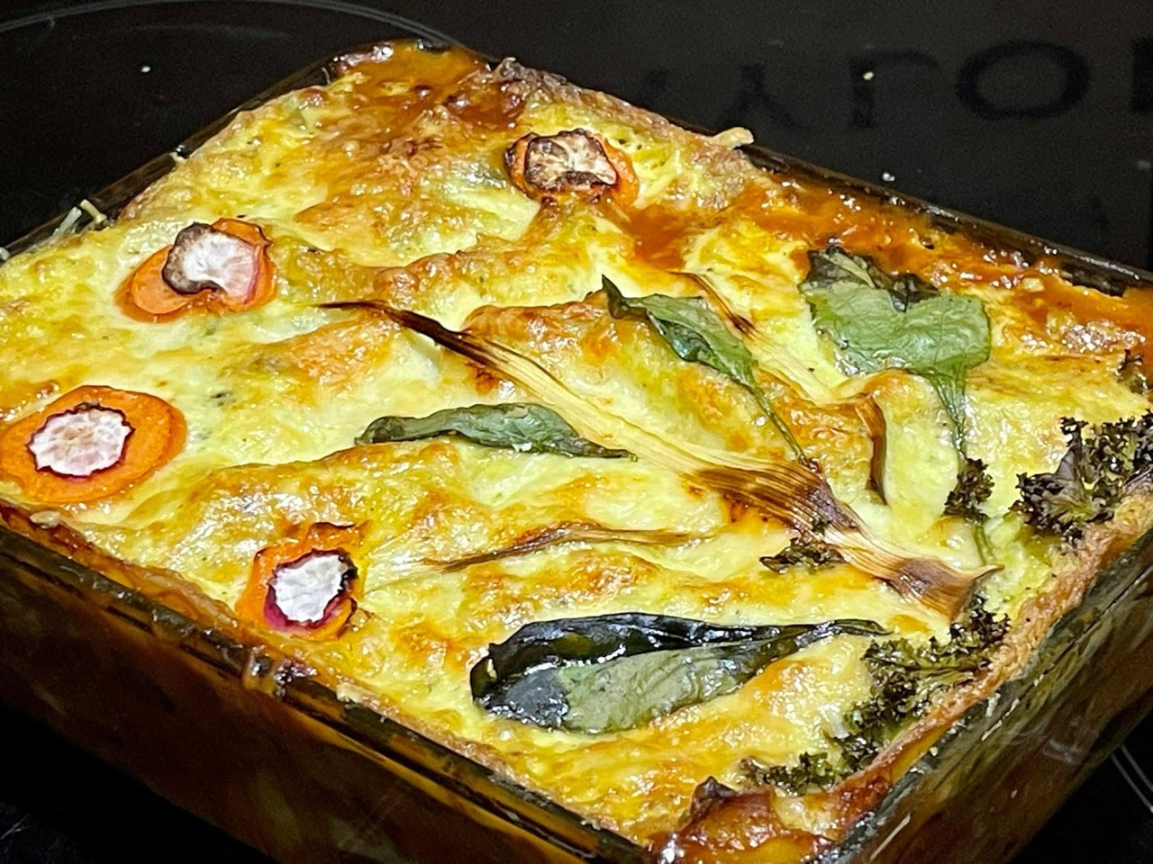 A baked vegan garden lasagna in a glass dish with golden topping, carrots, greens and herbs.