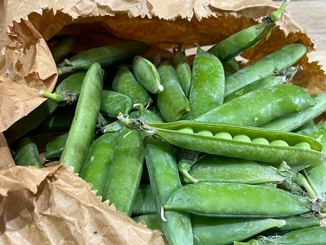 Fresh pea pods in a brown paper bag, with one pod opened to show peas.