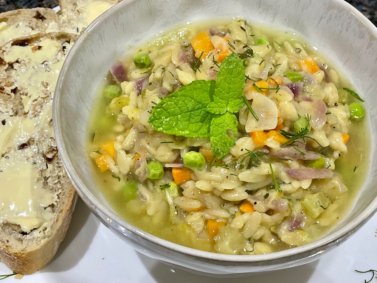 A bowl of minted pea and courgette orzo risotto with herbs, mint leaves and bread on the side.