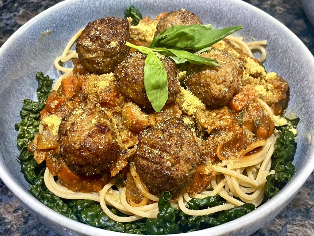 Vegan meatballs and spaghetti with tomato sauce, kale, basil and grated vegan cheese in a blue bowl.