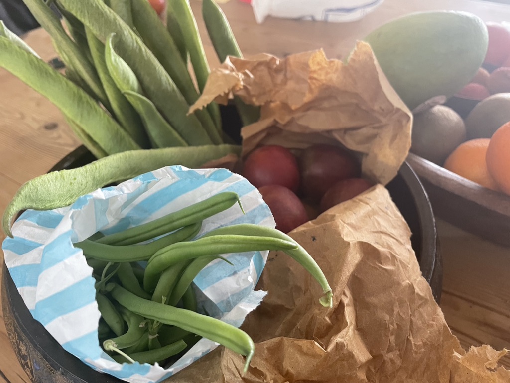 Green beans, plums and fruit gathered on a wooden table in paper bags and bowls.