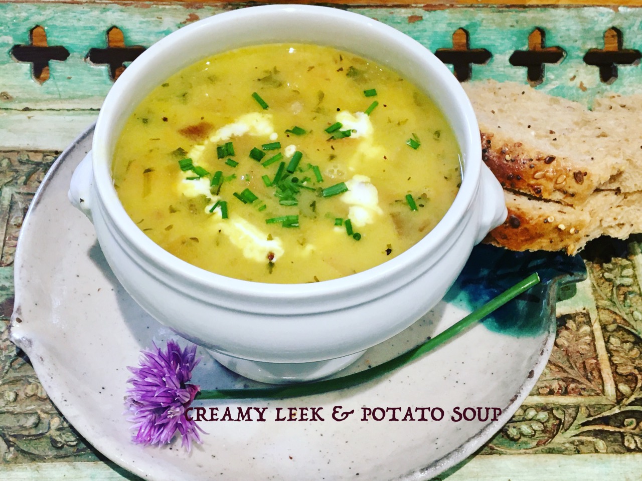 Creamy leek and potato soup in a white bowl with bread and chive flowers.
