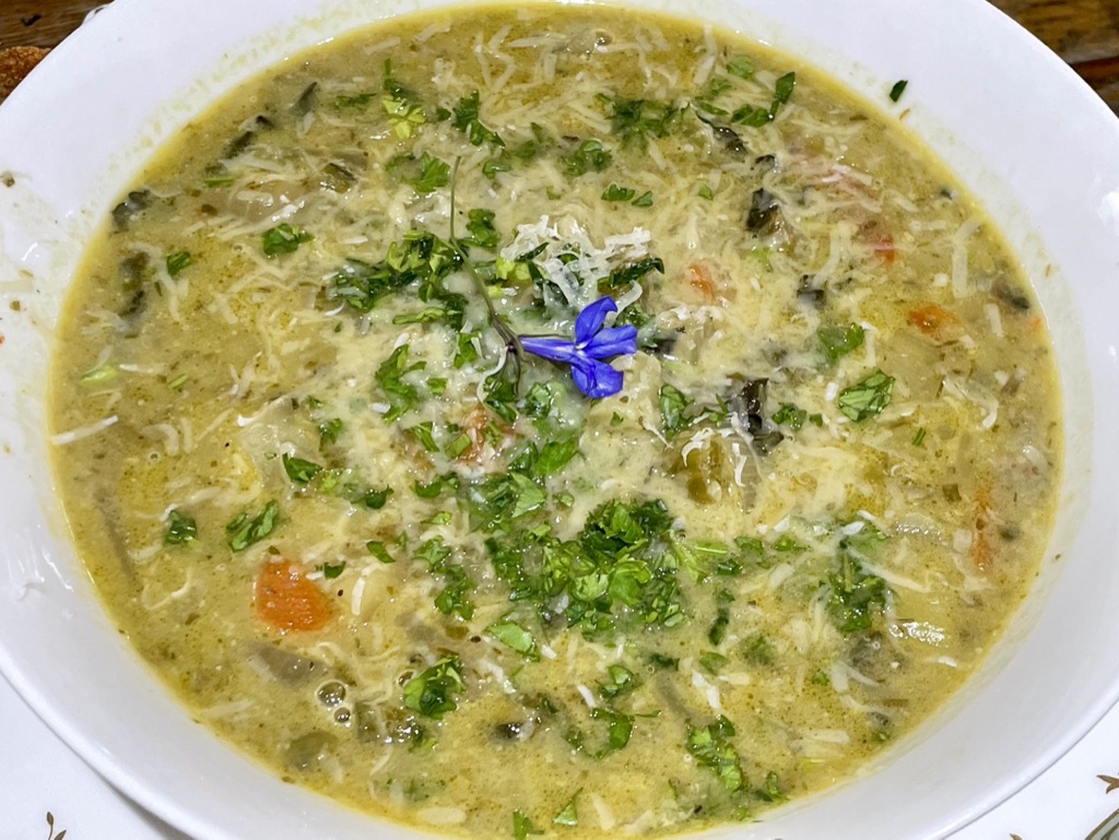Leek, potato and watercress soup topped with herbs, grated vegan cheese and a small blue edible flower.